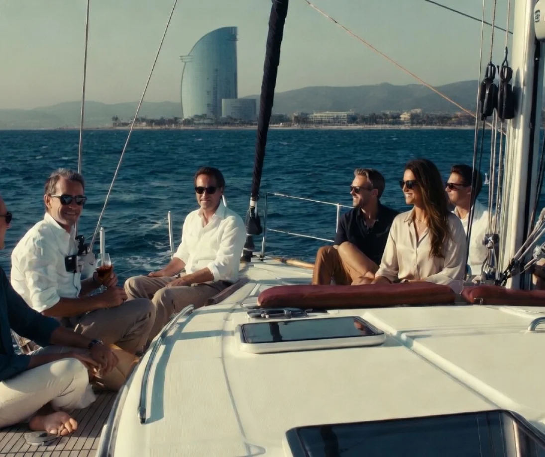 Tres personas hablando relajadas en un velero de lujo frente al skyline de Barcelona al atardecer