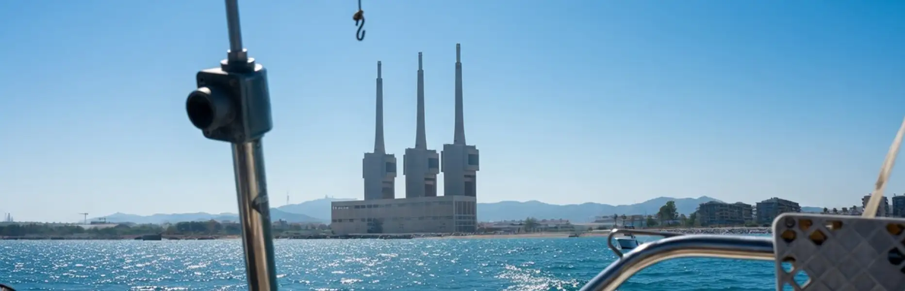 Vista des d’un veler navegant davant la costa de Barcelona amb el mar blau i les torres de la central de Sant Adrià al fo