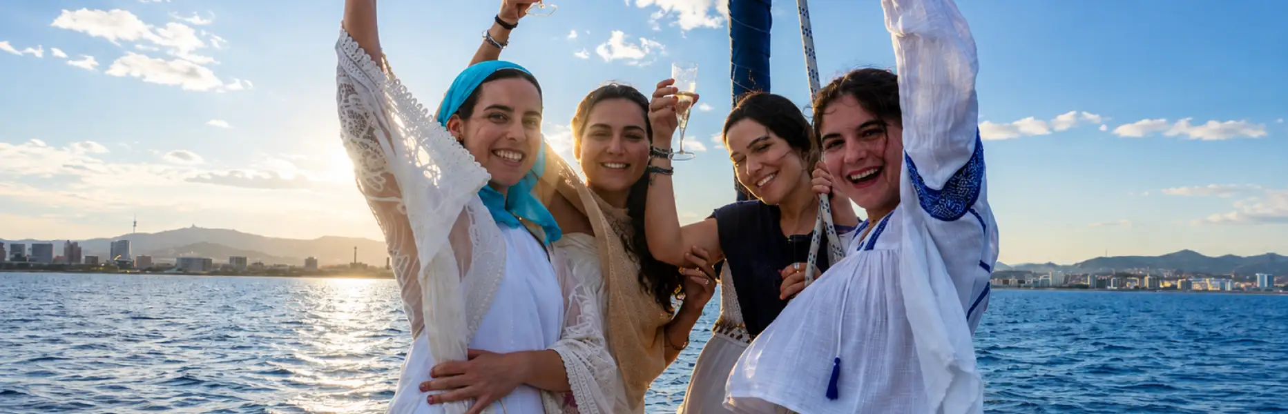 Gruppo di amiche che brindano con cava a prua di una barca a vela privata di fronte alla costa di Barcellona al tramonto