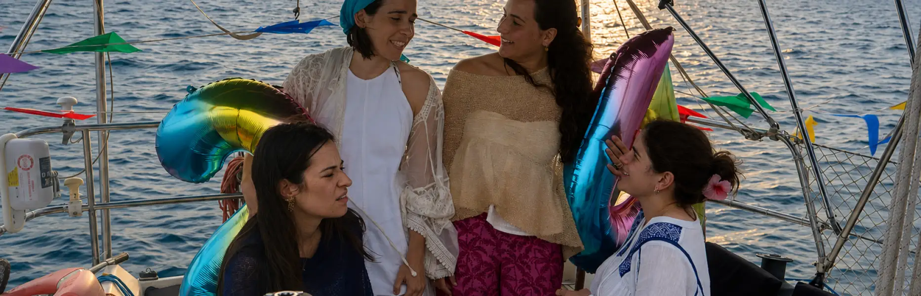 Gruppe von Freundinnen, die einen Geburtstag auf einer privaten Segelyacht vor der Küste von Barcelona bei Sonnenuntergang mit Luftballons und Dekoration feiern
