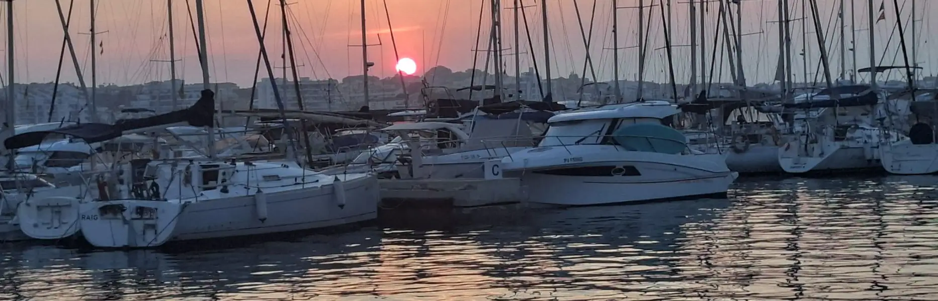 Sailing boat and yachts moored in a Barcelona marina at sunset with the sun on the horizon and a calm sea