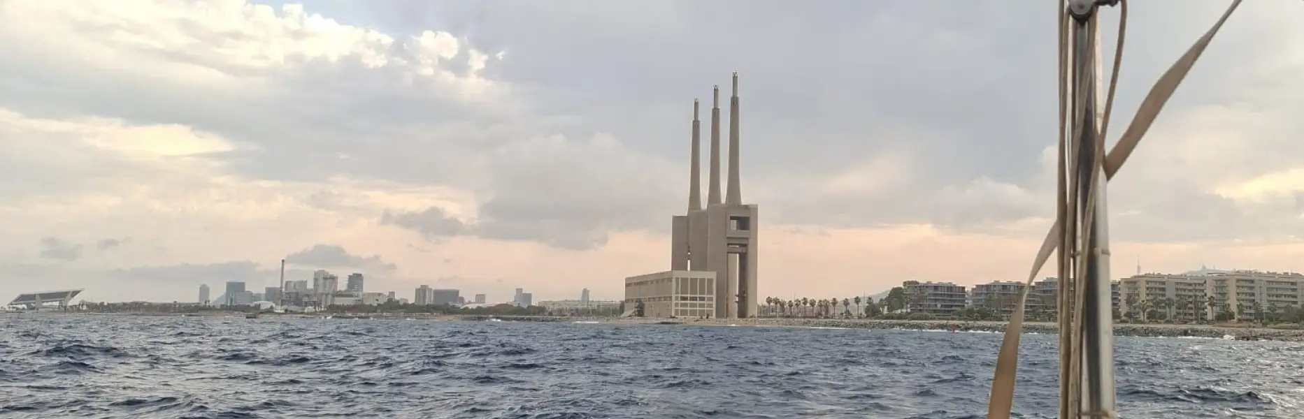 Blick von einem Segelboot, das vor der Küste von Barcelona segelt, mit leicht bewegter See und den drei Schornsteinen im Hintergrund