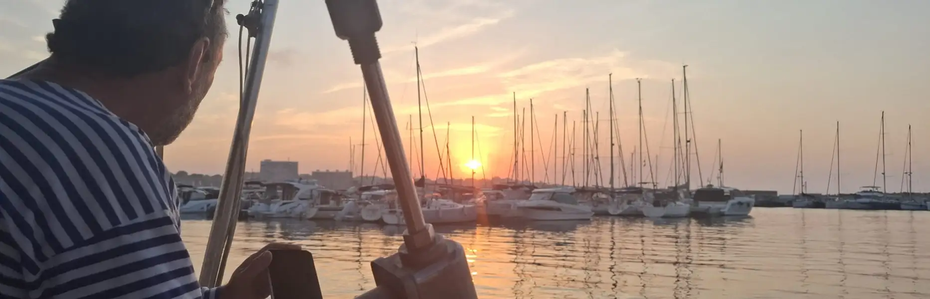 Patró en veler privat observant la posta de sol sobre el port esportiu de Barcelona amb nombrosos velers amarrats