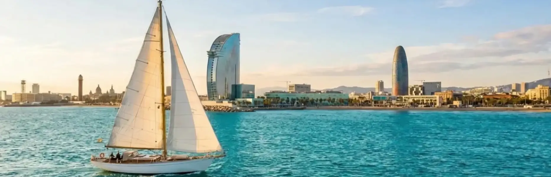 Velero de charter navegando al atardecer frente al skyline marítimo de Barcelona