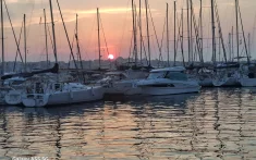 Veler i iots amarrats en un port de Barcelona al capvespre amb el sol sobre l’horitzó i el mar en calma